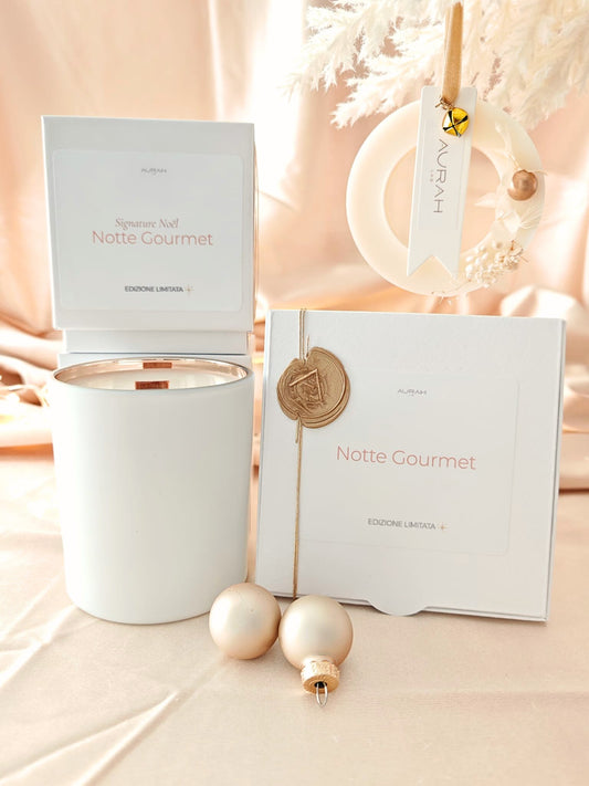Set Noël Notte Gourmet di Aurah Lab: candela bianca decorata con nappina oro, ghirlandina profumata e packaging coordinato, fotografati su un set luminoso beige natalizio con sfere decorative.
