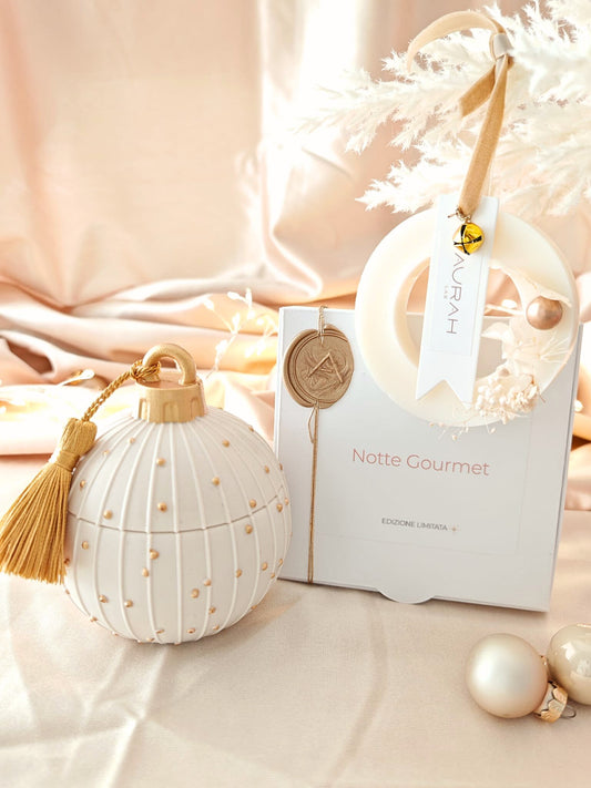 Set Noël Notte Gourmet di Aurah Lab: candela bianca decorata con nappina oro, ghirlandina profumata e packaging coordinato, fotografati su un set luminoso beige natalizio con sfere decorative.