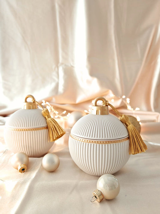 Candela decorativa Aurah Lab in forma di sfera bianca con dettagli dorati e nappina oro, fotografata su un set natalizio caldo con luci soffuse e sfere decorative.