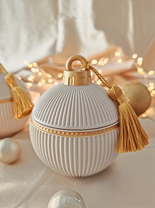 Candela decorativa Aurah Lab in forma di sfera bianca con dettagli dorati e nappina oro, fotografata su un set natalizio caldo con luci soffuse e sfere decorative.