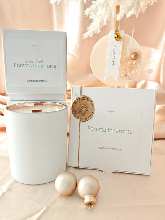Set Noël Foresta Incantata di Aurah Lab: candela bianca decorata con nappina oro, ghirlandina profumata e packaging coordinato, fotografati su un set luminoso beige natalizio con sfere decorative.