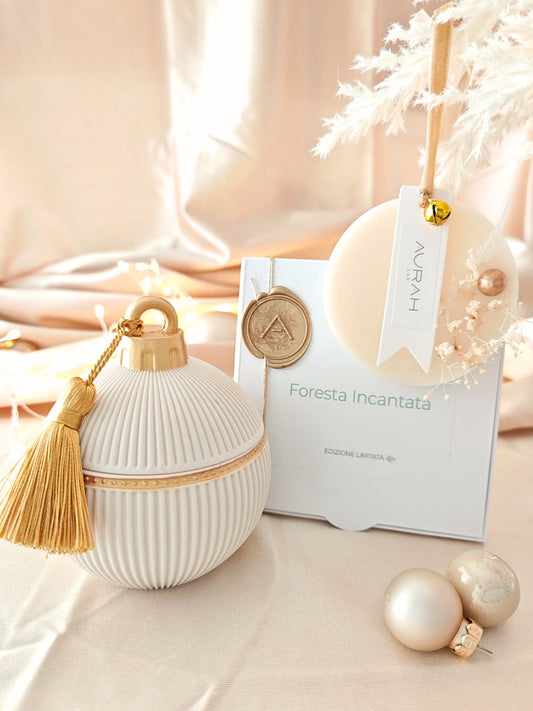 Set Noël Foresta Incantata di Aurah Lab: candela bianca decorata con nappina oro, ghirlandina profumata e packaging coordinato, fotografati su un set luminoso beige natalizio con sfere decorative.